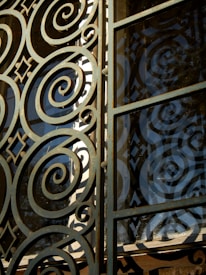 Intricate metalwork featuring spirals and geometric patterns integrated into a gate or window. The structure is crafted with ornate, swirling designs, casting shadows and reflections against a glass panel.