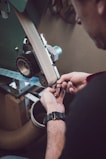 A person is working with a belt sander, carefully adjusting or inspecting the machine. The scene suggests a focus on craftsmanship or repair work, with a close-up view of the hands and the tool, highlighting the precision required.