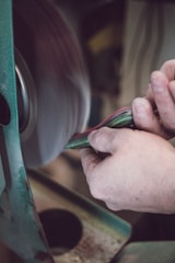 A person is using sandpaper on a rotating sanding machine to smooth an object. The hands are firmly holding multiple sheets of colorful material, and the machine is actively smoothing it. The scene captures a close-up view of the process, with focus on the hands and the edge of the sanding machine.