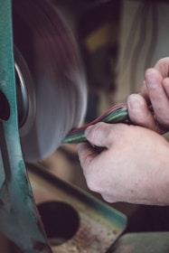 A person is using sandpaper on a rotating sanding machine to smooth an object. The hands are firmly holding multiple sheets of colorful material, and the machine is actively smoothing it. The scene captures a close-up view of the process, with focus on the hands and the edge of the sanding machine.