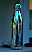 A metallic water bottle with a reflective surface that mirrors the surroundings, sitting on a wooden surface. Part of a wallet or pouch is visible next to the bottle, and there's a blurred indoor background.