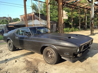 A vintage black muscle car is parked on a dirt lot next to a rusty industrial structure. The car has sleek lines and a matte finish, and it's positioned under a clear blue sky. In the background, there are suburban houses with trees providing greenery.