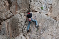 A person is climbing a vertical rock face, secured by ropes and climbing gear. The rocky surface is rugged and textured, displaying light and dark shades of gray and brown. The climber is wearing a black shirt, blue jeans, and a red harness with other climbing equipment attached.