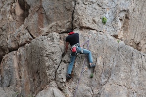 A person is climbing a vertical rock face, secured by ropes and climbing gear. The rocky surface is rugged and textured, displaying light and dark shades of gray and brown. The climber is wearing a black shirt, blue jeans, and a red harness with other climbing equipment attached.