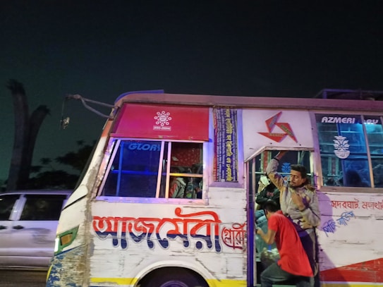 A bus with colorful decorations and signage is parked or moving slowly on a road at night. Several people are visible; one is hanging onto the door while another stands on the steps. The bus appears worn with visible paint peeling and damage.