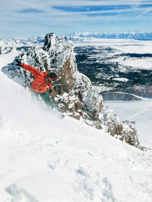 A snowboarder in a red jacket launching off a rock band above a frozen lake, with the snowy Eastern Sierra plateau behind