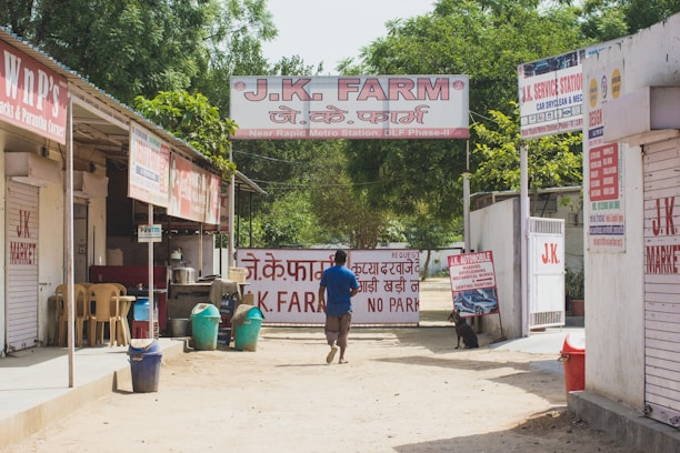 A person walks down a dirt path lined with small shops and service centers. The shops have signs in both English and Hindi, with one prominently displaying 'J.K. Farm' above the entrance. There are several plastic chairs and trash bins in front of one shop. Lush green trees are visible in the background, adding a touch of nature to the urban setting.