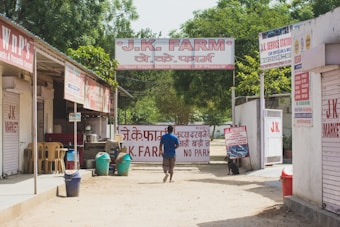 A person walks down a dirt path lined with small shops and service centers. The shops have signs in both English and Hindi, with one prominently displaying 'J.K. Farm' above the entrance. There are several plastic chairs and trash bins in front of one shop. Lush green trees are visible in the background, adding a touch of nature to the urban setting.