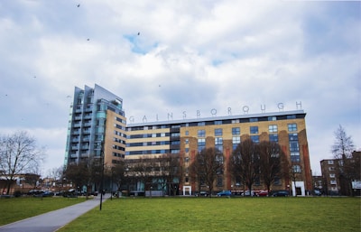 A large multi-story building with modern and older architectural elements, featuring a prominent sign on the roof spelling out 'GAINSBOROUGH'. The building is surrounded by trees and a grassy area with a paved walkway. A few birds are flying in the cloudy sky.