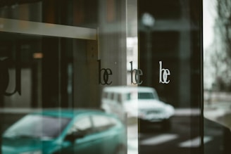 Reflections of a street scene are seen on a glossy surface, with the word 'be' repeated multiple times on the glass. The image includes the blurry forms of parked cars outside and an urban setting with a muted, overcast atmosphere.