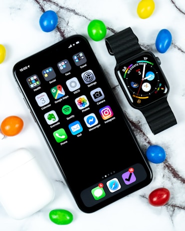A smartphone and a smartwatch are placed on a marble surface surrounded by colorful candy-coated chocolates. The phone displays various app icons on the home screen, while the smartwatch shows digital time and other information. Nearby, a white wireless earbuds case is visible.