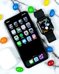 A smartphone and a smartwatch are placed on a marble surface surrounded by colorful candy-coated chocolates. The phone displays various app icons on the home screen, while the smartwatch shows digital time and other information. Nearby, a white wireless earbuds case is visible.