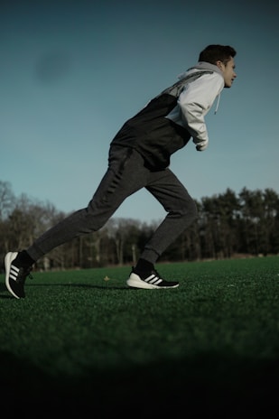 A person is captured mid-stride, running across a field. They are wearing a two-tone jacket, dark track pants, and black sneakers with white soles on a grass surface. The background consists of trees under a cloudy sky, suggesting an outdoor or park setting.