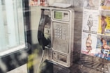 A vintage public payphone with a corded handset is mounted on a wall inside a phone booth. The metallic casing of the phone is slightly worn and aged. Several colorful advertisements with images are posted around the phone, promoting various services.