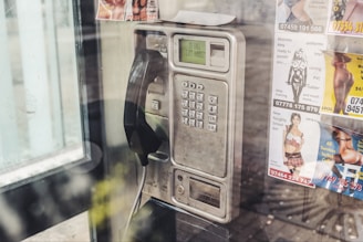 A vintage public payphone with a corded handset is mounted on a wall inside a phone booth. The metallic casing of the phone is slightly worn and aged. Several colorful advertisements with images are posted around the phone, promoting various services.