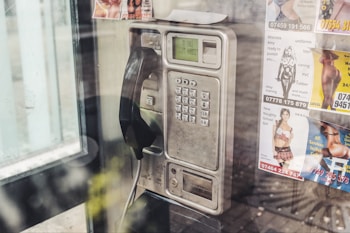 A vintage public payphone with a corded handset is mounted on a wall inside a phone booth. The metallic casing of the phone is slightly worn and aged. Several colorful advertisements with images are posted around the phone, promoting various services.