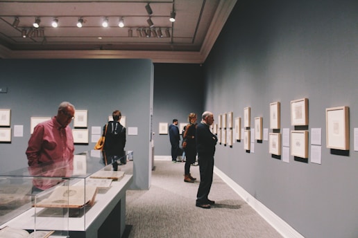 A group of people are observing framed artworks on the walls of an art gallery. The room has dim lighting with spotlights on the ceiling, highlighting the artwork. There is a display case containing objects or documents on the left side.