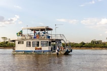 A houseboat is floating on a calm river with people visible on board, some seated on the upper deck. The boat has a flag attached, chairs, and tables on the top, with jungle or forestation visible in the background and a sunny, clear sky above. The boat has an open structure above the deck with railings and life preservers hanging. Two individuals are seen at the front of the boat, one holding a rope.
