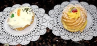Two pastries are placed on decorative white lace doilies. The left pastry has a light frosting with an orange slice, chopped nuts, and a small green piece of candied fruit. The right pastry is circular and layered, topped with yellow cream, powdered sugar, a red berry, and nuts.