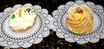 Two pastries are placed on decorative white lace doilies. The left pastry has a light frosting with an orange slice, chopped nuts, and a small green piece of candied fruit. The right pastry is circular and layered, topped with yellow cream, powdered sugar, a red berry, and nuts.