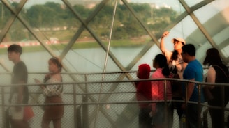 A group of people stands on a fenced platform inside a large glass structure. One person, wearing a cap, raises a smartphone, perhaps taking a photo or selfie. The background shows a serene outdoor landscape with greenery and a body of water visible through the geometric glass panels.
