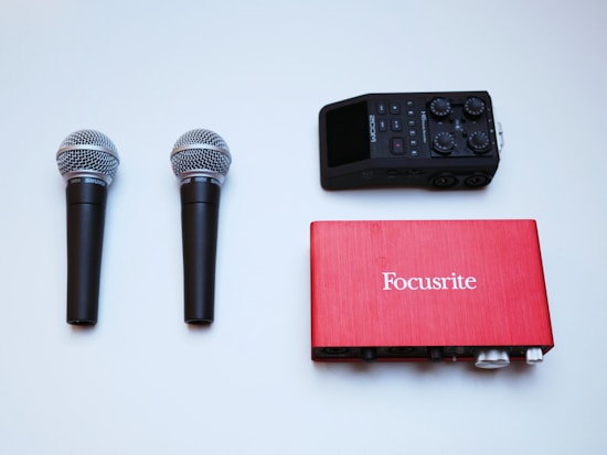 Two microphones with black handles and silver mesh heads, a red Focusrite audio interface, and a black Zoom audio recorder are neatly arranged on a flat, light-colored surface.