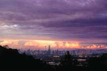 A panoramic view of a city skyline during sunset, with towering skyscrapers against a backdrop of dramatic clouds. The sky is a blend of purple and orange hues, with the silhouette of a hilly landscape framing the city.