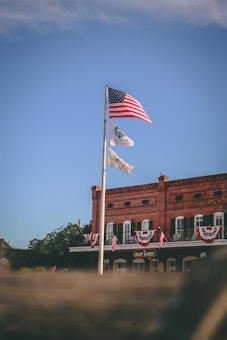 A historic brick building with white trim is adorned with patriotic banners and American flags. A tall flagpole holds the American flag, alongside other flags. The sky is clear and blue, providing a serene backdrop.