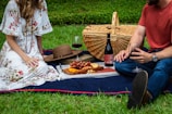 man and woman sitting on blue textile beside brown wicker picnic basket