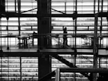A black and white image of a modern architectural space with large windows and metal beams. Two people are visible: one sitting at a table and the other standing, silhouetted against the bright light from the windows. The design emphasizes horizontal and vertical lines, creating a structured and geometric appearance.