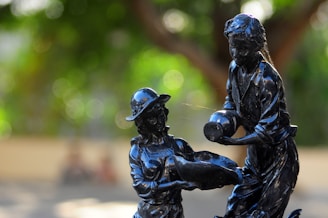 A pair of dark statues depicting two figures, one holding a jug and the other holding a basin, against a blurred background with vibrant green, brown, and bokeh highlights.