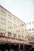 A large building with multiple floors features rows of windows and a sign that reads 'City Liquidators.com'. The exterior of the building is adorned with numerous colorful triangular flags strung across horizontally. Below, a storefront area shows displays and balloons, suggesting a festive or market-like environment.