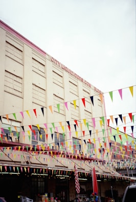 A large building with multiple floors features rows of windows and a sign that reads 'City Liquidators.com'. The exterior of the building is adorned with numerous colorful triangular flags strung across horizontally. Below, a storefront area shows displays and balloons, suggesting a festive or market-like environment.