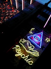 A neon sign with a glowing eye inside a triangle, accompanied by a star and crescent moon motif. Below, the word 'psychic' is illuminated in bright yellow neon. The scene is set against a backdrop of reflective and textured surfaces.