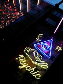 A neon sign with a glowing eye inside a triangle, accompanied by a star and crescent moon motif. Below, the word 'psychic' is illuminated in bright yellow neon. The scene is set against a backdrop of reflective and textured surfaces.