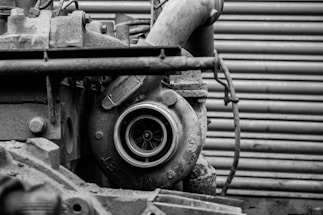 A close-up view of an industrial machine component, possibly a turbocharger or similar machinery part. The metal surfaces appear weathered, with visible wear, rust, and texture. The background consists of horizontal, corrugated metal resembling a garage door or industrial wall.