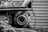 A close-up view of an industrial machine component, possibly a turbocharger or similar machinery part. The metal surfaces appear weathered, with visible wear, rust, and texture. The background consists of horizontal, corrugated metal resembling a garage door or industrial wall.
