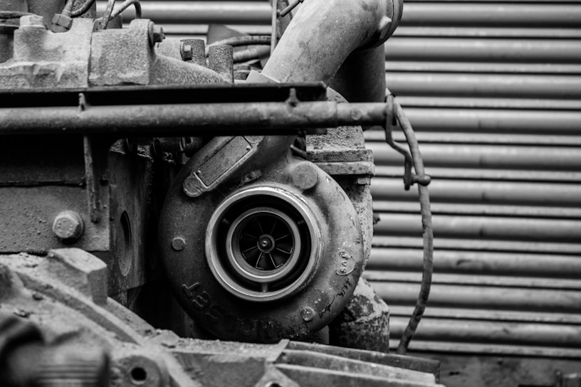 A close-up view of an industrial machine component, possibly a turbocharger or similar machinery part. The metal surfaces appear weathered, with visible wear, rust, and texture. The background consists of horizontal, corrugated metal resembling a garage door or industrial wall.