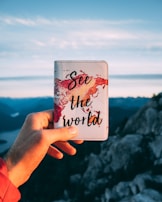A hand holds a passport cover with a watercolor world map design and the words 'See the world' written on it. The background features a scenic mountain view with a partially cloudy sky.