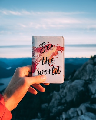 A hand holds a passport cover with a watercolor world map design and the words 'See the world' written on it. The background features a scenic mountain view with a partially cloudy sky.