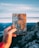 A hand is holding a journal with a world map design and the text 'I haven't been everywhere, but it's on my list.' The background features a scenic view of mountains and a sky with clouds.