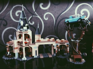 A LEGO model of a castle with tall towers and intricate details. A blue vintage car is perched on a tree, showing an imaginative assembly. The background features a gray wall with white swirling patterns.