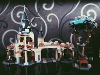A LEGO model of a castle with tall towers and intricate details. A blue vintage car is perched on a tree, showing an imaginative assembly. The background features a gray wall with white swirling patterns.