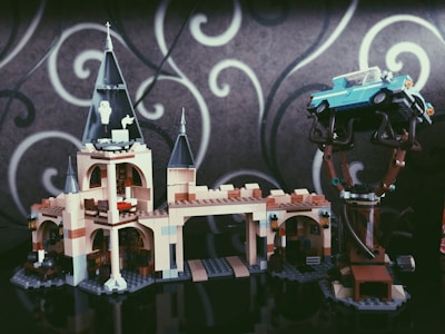 A LEGO model of a castle with tall towers and intricate details. A blue vintage car is perched on a tree, showing an imaginative assembly. The background features a gray wall with white swirling patterns.