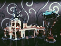 A LEGO model of a castle with tall towers and intricate details. A blue vintage car is perched on a tree, showing an imaginative assembly. The background features a gray wall with white swirling patterns.