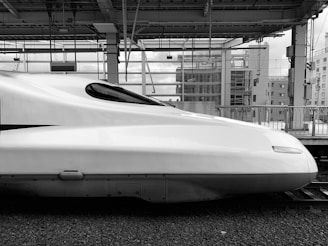 A sleek, modern-looking high-speed train positioned at a station platform. The train's aerodynamic design is emphasized by its smooth, elongated front section. The background shows industrial and urban elements with metal beams, glass structures, and buildings.