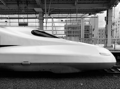 A sleek, modern-looking high-speed train positioned at a station platform. The train's aerodynamic design is emphasized by its smooth, elongated front section. The background shows industrial and urban elements with metal beams, glass structures, and buildings.