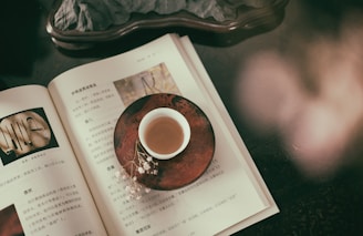 An open book with text and images rests on a dark surface. A small cup filled with tea sits on a decorative saucer on top of the book. There are delicate white flowers next to the cup, adding to the serene and contemplative atmosphere.