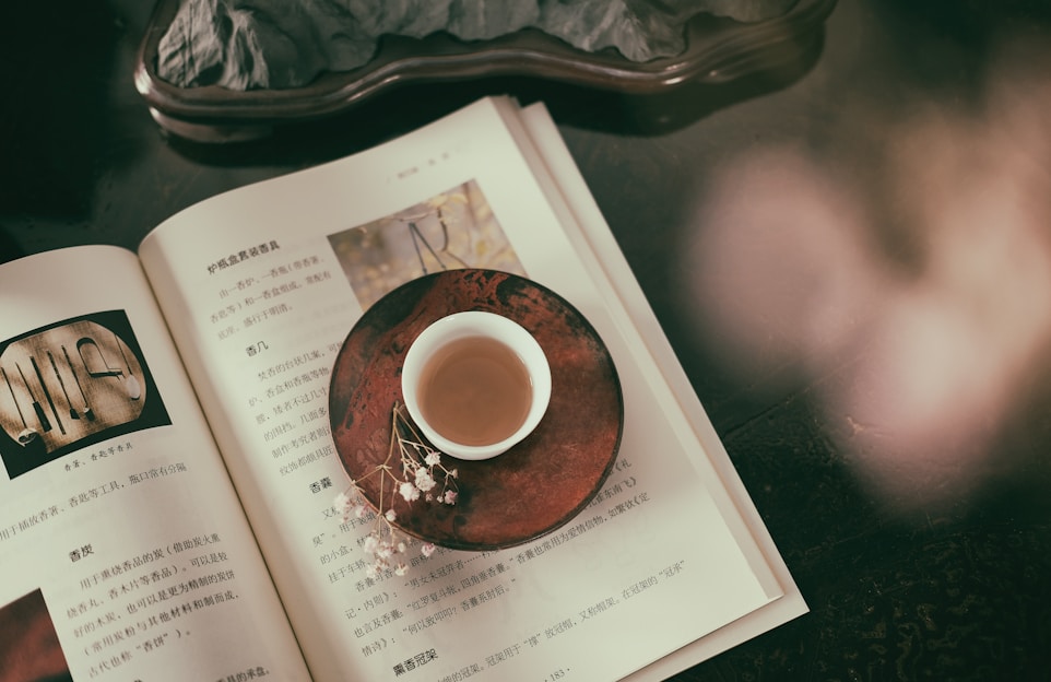 An open book with text and images rests on a dark surface. A small cup filled with tea sits on a decorative saucer on top of the book. There are delicate white flowers next to the cup, adding to the serene and contemplative atmosphere.