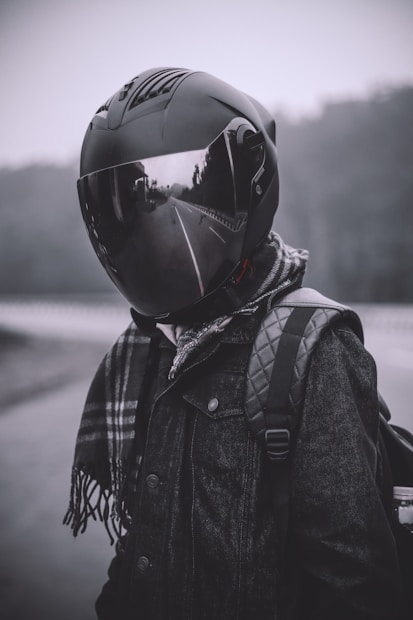 A person wearing a large, reflective motorcycle helmet and a dark coat stands on a misty road. The helmet reflects the surrounding scenery, including trees and an empty road. A checkered scarf is draped over the coat, and a backpack is worn over one shoulder.